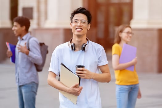 College Life. Portrait of asian student in glasses holding coffee and textbook, his classmates talking in background photo