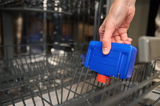 Close-up of hand holding blue dishwasher cleaner bottle and placing it inside empty dishwasher. Concept of appliance maintenance and cleaning routine photo