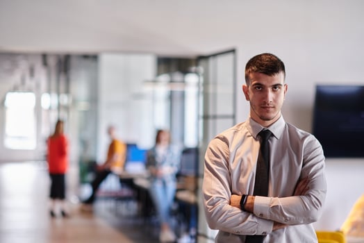 A young business leader stands with crossed arms in a modern office hallway, radiating confidence and a sense of purpose, embodying a dynamic and inspirational presence. photo