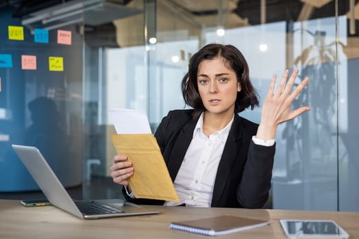 Young businesswoman holding envelope in modern office, expressing confusion while discussing financial documents. Laptop and smartphone visible, creating sense of corporate frustration. photo