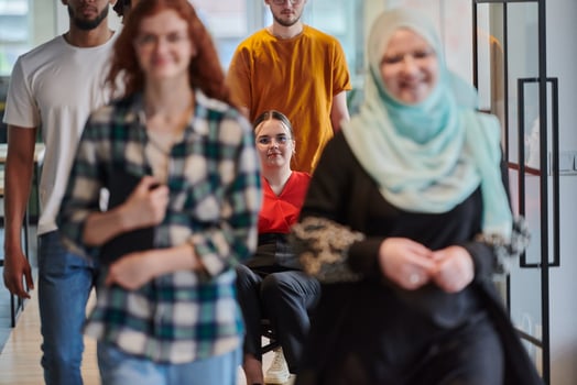 A diverse group of business people walking a corridor in the glass-enclosed office of a modern startup, including a person in a wheelchair and a woman wearing a hijab, showing a dynamic mix of photo
