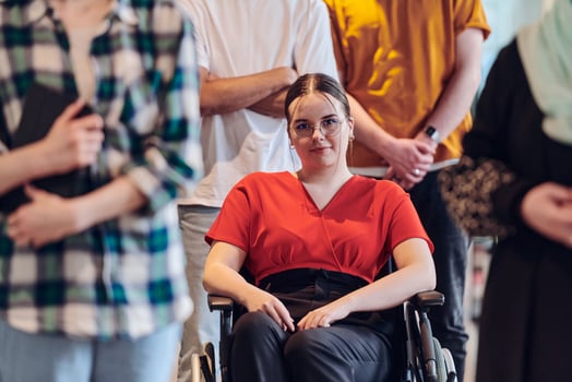 A diverse group of business people walking a corridor in the glass-enclosed office of a modern startup, including a person in a wheelchair and a woman wearing a hijab, showing a dynamic mix of photo