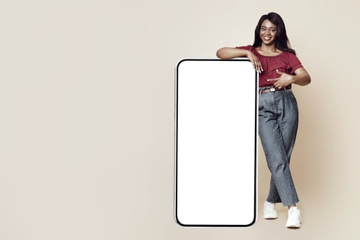 Stylish millennial black woman pointing at big mobile phone with white blank display, mockup, happy african american lady in casual recommending newest mobile application, beige studio background photo
