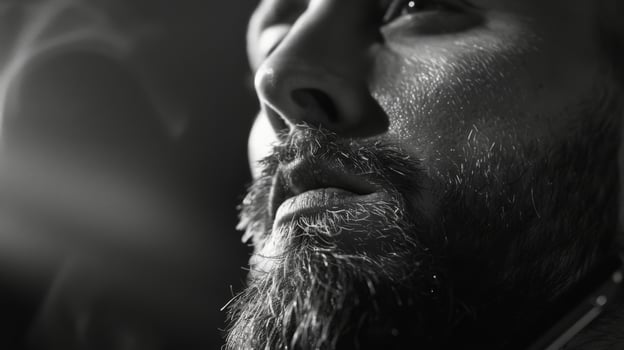 A man with a well groomed beard gazes thoughtfully with a serious expression, illuminated by soft lighting, in a relaxed and artistic environment during the evening. photo