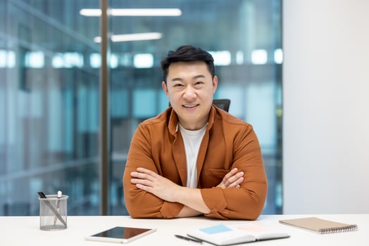 Asian businessman is confidently sitting at his desk in a modern office with his arms crossed, smiling warmly at the camera photo