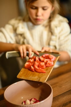 A young girl in an apron slices a tomato for a salad. Fresh vegetables are on the table, and she watches her cutting closely. photo