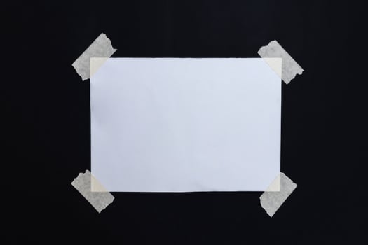 Blank note paper for writing a message attached with tape on all four sides on a black background photo