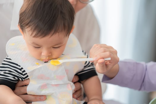 Baby enjoying mealtime as caregiver feeds with a spoon in a bright indoor setting photo