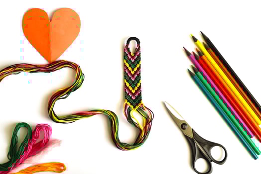 Unfinished woven friendship bracelet with the skeins of thread, scissors, cut heart and selected palette of crayons colors on white background photo