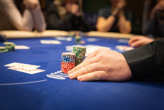 A player's hand rests on poker chips on a blue felt table with visible playing cards, including aces, and blurred players in the background. photo