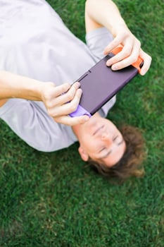 Joyful man smiling and having fun while playing games with portable console and lying on the grass outdoors. Technology and game addiction concept photo