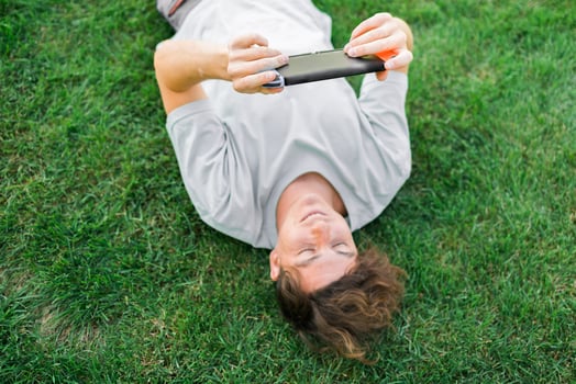Joyful man smiling and having fun while playing games with portable console and lying on the grass outdoors. Technology and game addiction concept photo