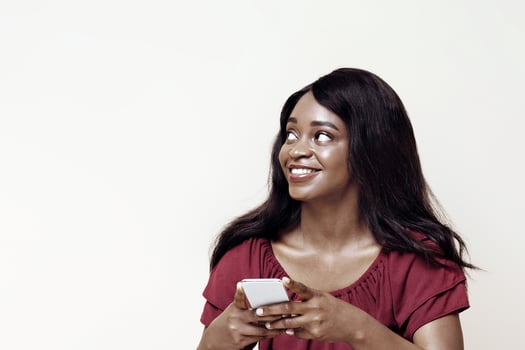 Pretty long-haired young black woman with newest cellphone sitting on floor, looking at copy space over beige studio background, using nice dating, entertaining mobile app on smartphone, panorama photo