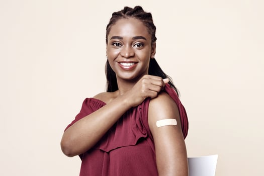 Happy black lady sitting on chair, showing adhesive medical band on her shoulder after vaccination and smiling at camera, beige studio background, copy space. Immunization during COVID-19 pandemic photo