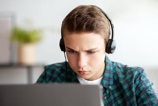 Concentrated teenager using laptop, wearing headset, sitting at home alone, copy space photo