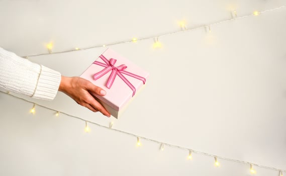 Pink gift box with a bow in female hand in white sweater on a light background. Christmas garlands on the wall photo