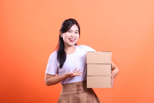 Cheerful woman with dark hair and a white shirt, holds a stack of three cardboard boxes and presenting with hand. isolated on orange background photo