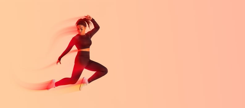 Fitness lifestyle. Motivated African American lady jumping in air over peach neon studio background, athletic woman making jump exercises, enjoying cardio workout, full length, copy space photo