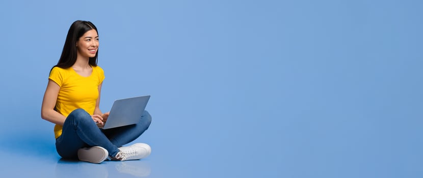 Smiling young asian woman sitting on floor with laptop computer over purple studio background, looking away at copy space, millennial korean lady studying or working with notebook photo