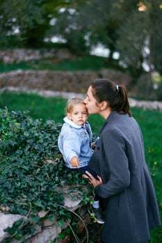 Mom kisses the temple of a little girl sitting on a fence in the park photo