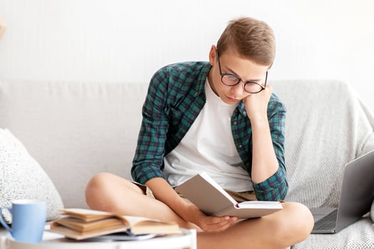 Tired teenager studying at home, sitting on couch and reading book, e-education concept, free space photo