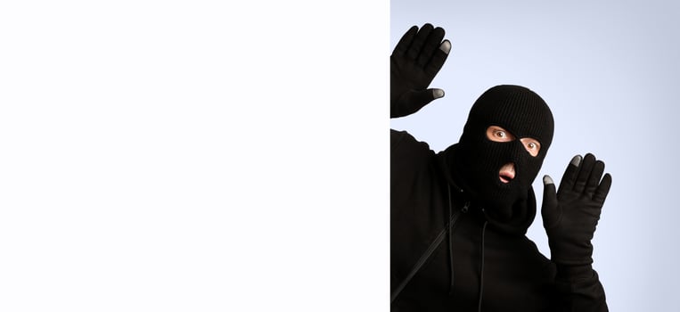 Scared thief in a balaclava and gloves raising hands up and looking out of blank sign with copyspace for your text photo