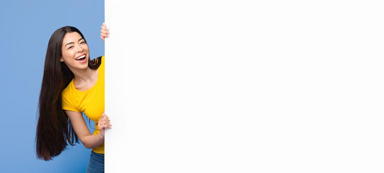 Cheerful asian girl looking out of blank white advertisement sign board with free space, standing over purple background, panorama photo