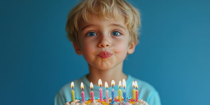 Young boy with blue eyes prepares to blow out birthday candles on a colorful cake against a blue background photo