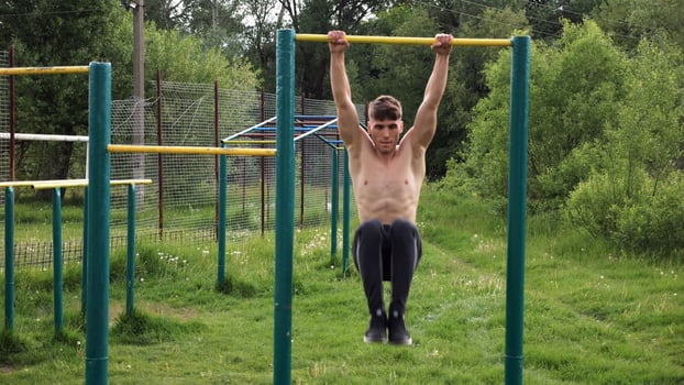Strong guy training abdominal muscles on horizontal bar at sports ground. Young sportsman exercising on pull-up bar outdoor. Sporty athlete working out at nature. Concept of sportive lifestyle photo