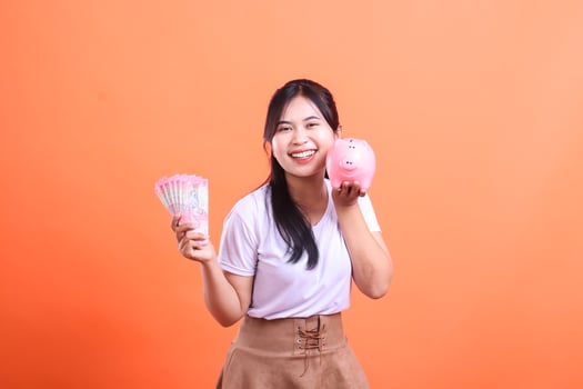 Smiling young woman holding cash and a piggy bank. Saving money for the future and investment. isolated on orange background photo