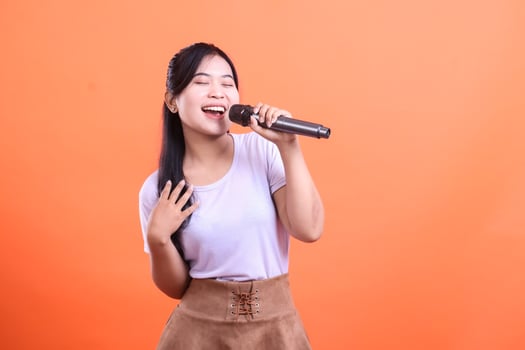 Enthusiastic young woman passionately singing into a microphone, showcasing her vocal talent and joy, isolated on orange background photo