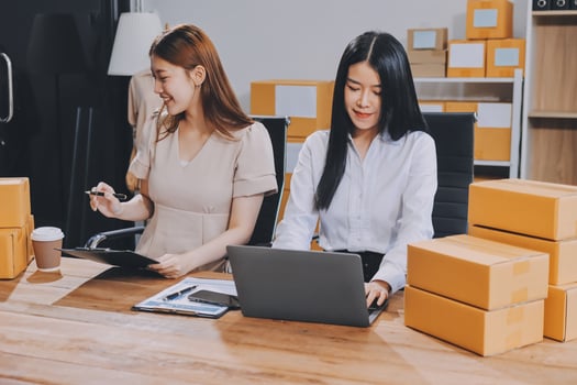 Two women sitting at a desk with boxes and laptops photo