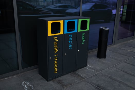 Three trash cans are lined up on a sidewalk, with one of them being green. The other two are blue and yellow. The trash cans are placed near a brick wall and a lighthouse photo
