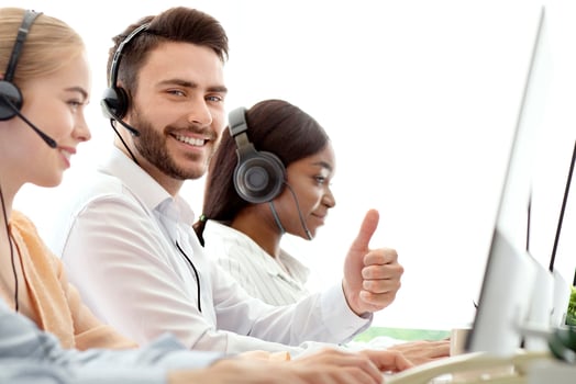 Happy male call centre operator showing thumb up gesture, working with his colleages at modern office. Positive tech support consultant providing telemarketing services photo