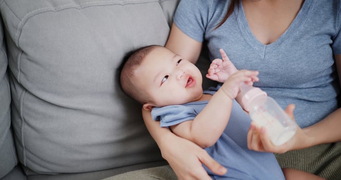 Selective focus shot, Beautiful mother gently holds her little son in her arms and feeds him with a bottle while sitting on a sofa in the living room, with an atmosphere full of love and warmth. photo