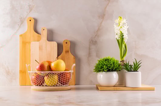 Chic kitchen still life with a set of cutting wooden boards, indoor flowering plants and a wicker fruit bowl on a marble beige countertop. Front view. photo