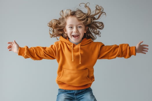 A joyful young girl with curly hair wearing an orange hoodie jumps in excitement against a neutral gray background. photo