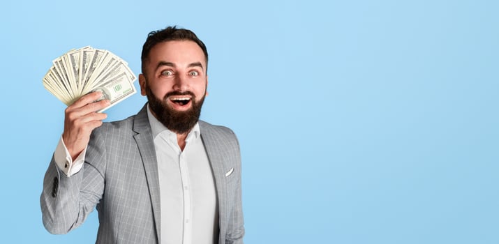 Cheerful bearded businessman in suit holding fan of dollar bills over blue background photo