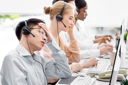 Overworked male hotline agent sleeping in front of computer monitor at call centre office, copy space. Exhausted Asian customer support manager napping at workplace near his colleagues photo
