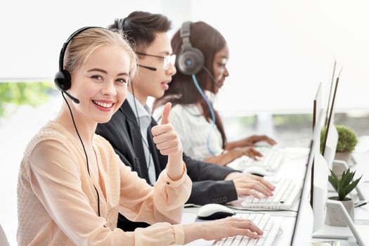 Happy female call centre consultant showing thumb up gesture and her colleages at modern office. Positive helpdesk operator working in telemarketing or in customer support service photo