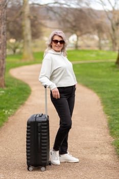Woman in sunglasses stands with a suitcase on a park alley, facing the camera. photo