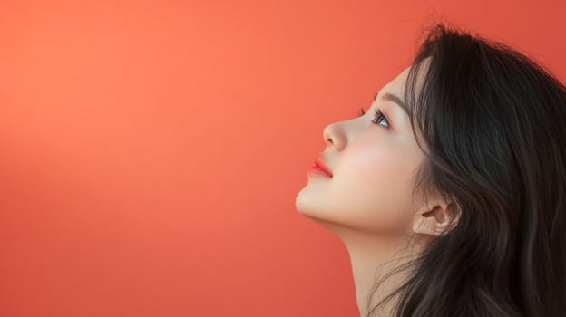 A young woman with soft, flowing dark hair looks upwards against a vibrant coral backdrop, showcasing beauty and serene contemplation. photo