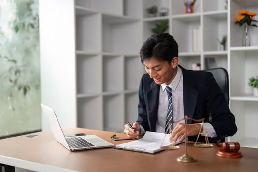 Young professional in suit smiling while taking notes at desk, working on laptop in modern office environment. photo