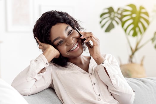 Mobile Communication. Cheerful african american woman talking on cellphone with friends while relaxing on comfortable couch in living room at home, sincerely smiling, having pleasant conversation photo