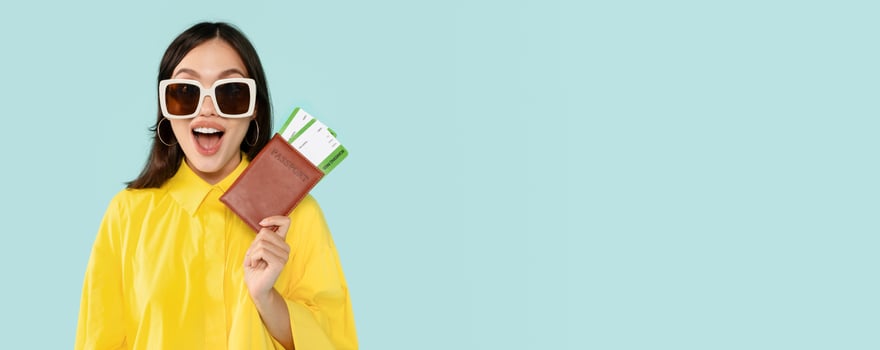 Vacation, travelling. Emotional young lady tourist wearing yellow shirt and sunglasses showing passport and flight tickets boarding pass, grimacing, blue studio background, panorama with copy space photo