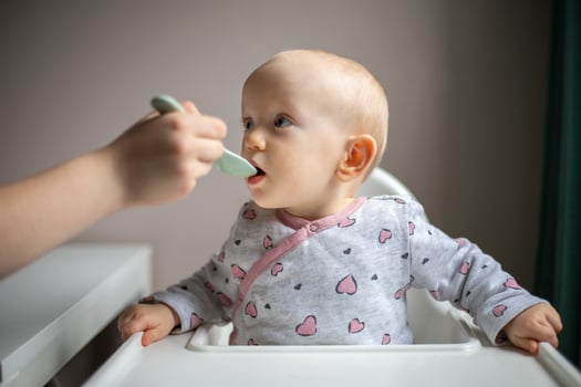 Baby girl eating blend mashed food sitting, on high chair, mother feeding child, hand with spoon for vegetable lunch, baby weaning, first solid food for young kid photo