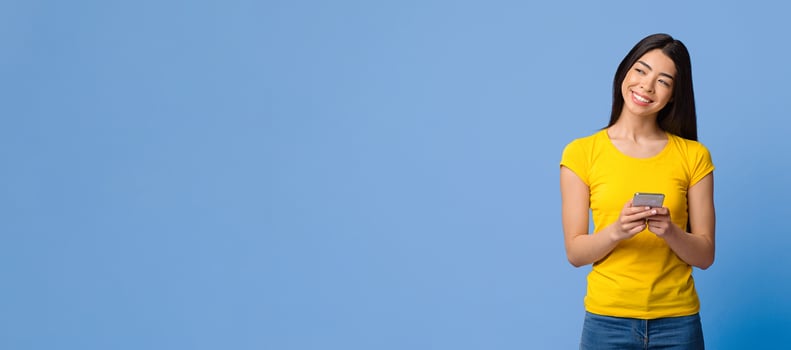 Dreamy Korean Woman Holding Smartphone In Hands And Looking Away At Copy Space On Purple Studio Background, Thinking About Mobile Offer And Online Sales, Chatting With Friends On Cellphone And Smiling photo