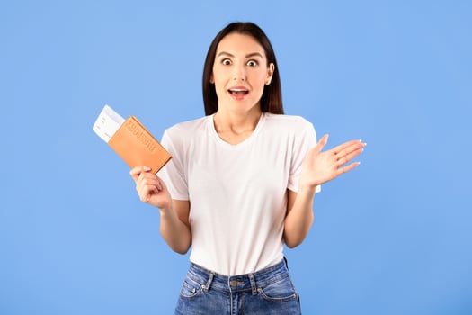Cheap Flights Concept. Portrait of surprised shocked woman holding passport and boarding pass ticket, standing isolated on blue studio background. Excited lady going on vacation after lockdown photo