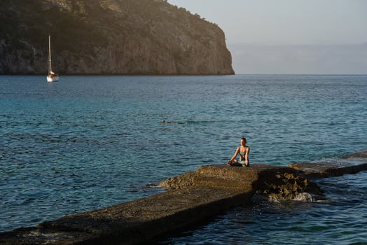 Woman sitting on boulder among blue ocean and taking Padmasana p photo
