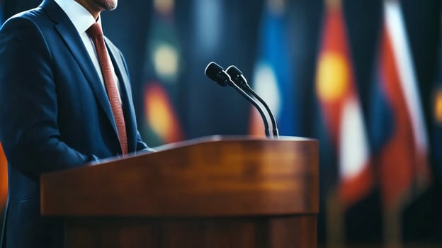 A speaker stands confidently at a podium during an international conference, with colorful flags representing various nations displayed behind. photo
